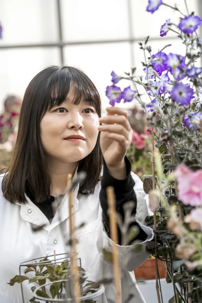 Een vrouw met donker haar op schouderlengte, gekleed in een witte laboratoriumjas, onderzoekt paarse en roze bloemen in een kas. Ze raakt zachtjes een paarse bloem aan terwijl ze omringd wordt door verschillende andere bloeiende planten. De achtergrond toont meer planten en diffuus natuurlijk licht.