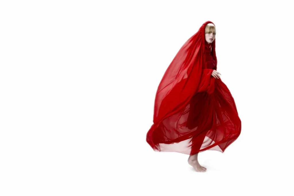 Een persoon staat blootsvoets tegen een witte achtergrond, gehuld in een golvende, doorschijnende rode stof die zijn hoofd bedekt en om zijn lichaam drapeert. Het figuur draagt een bijpassende rode outfit onder de stof. De pose straalt een gevoel van beweging uit, alsof ze midden in de stap staan: een creatieve knipoog naar bedrijfsfotografie.