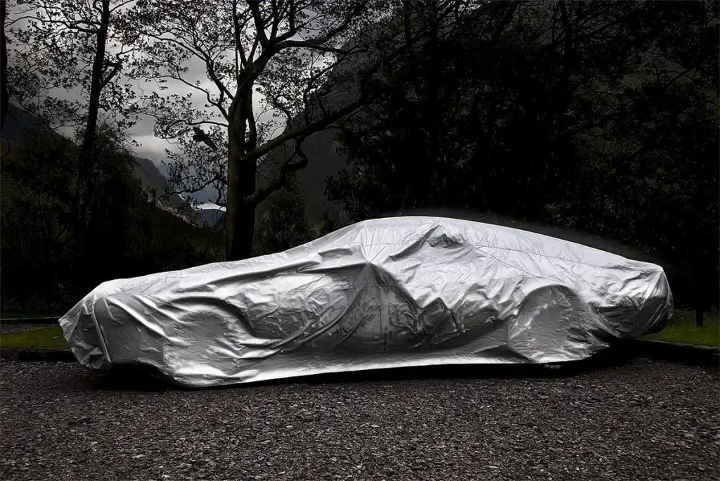 Een auto bedekt met een zilverkleurig zeildoek staat geparkeerd op een grindgebied. De achtergrond bestaat uit hoge, lommerrijke bomen en een bewolkte hemel, terwijl bergen in de verte gedeeltelijk zichtbaar zijn door het gebladerte. De setting lijkt een afgelegen, bergachtig gebied te zijn, perfect voor het vastleggen van serene bedrijfsfotografie.