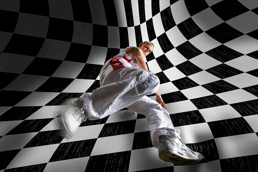 Een persoon in witte kleding en sneakers neemt een dynamische pose aan tegen een zwart-wit geblokte, gebogen achtergrond. Het perspectief is van onderaf, waardoor het lijkt alsof het individu klimt of zweeft. De verlichting benadrukt de kromming en het geometrische patroon van de achtergrond, wat doet denken aan creatieve bedrijfsfotografie.