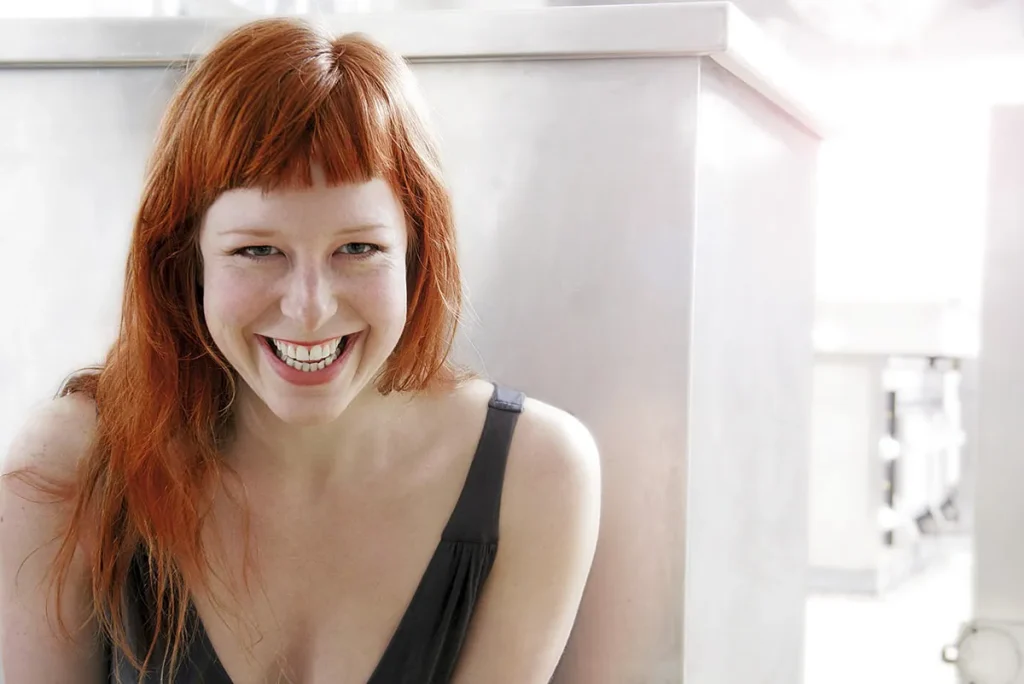 Een vrouw met lang rood haar en een pony lacht vrolijk en laat haar tanden zien. Ze draagt een donker, mouwloos topje. De achtergrond is helder, met lichtgekleurde, onscherpe elementen die een moderne binnenomgeving suggereren die vaak te zien is in bedrijfsfotografie. Haar vrolijke uitdrukking is het middelpunt van het beeld.