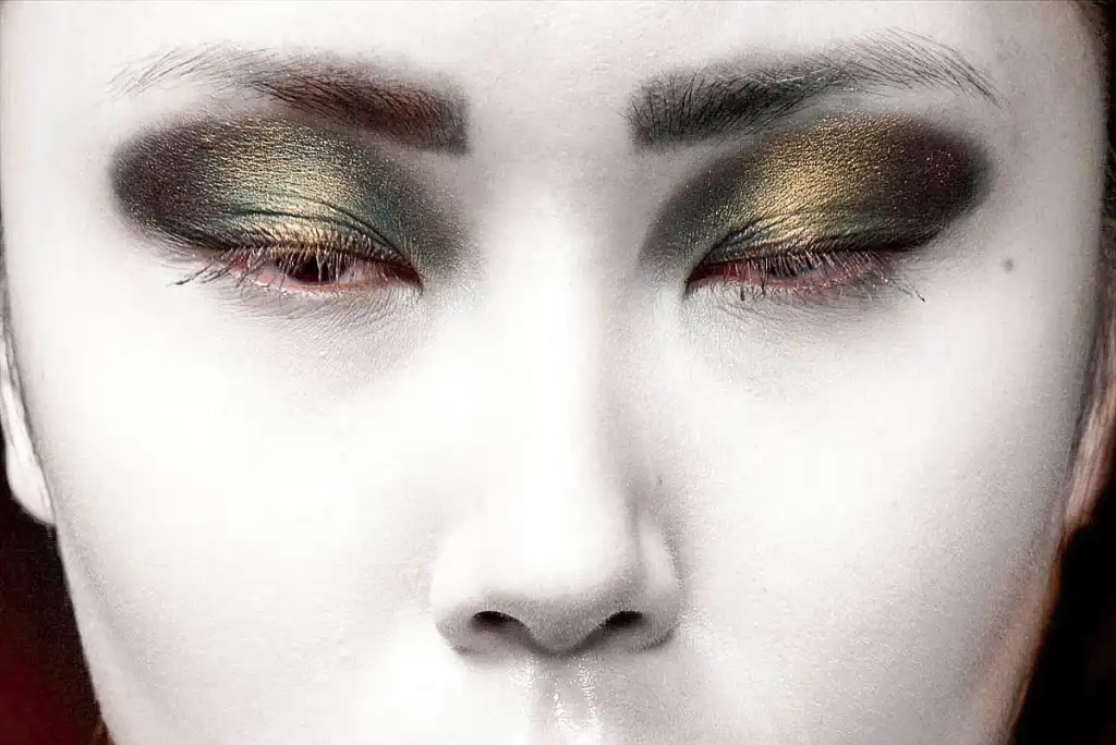 Close-up van iemands gezicht met gesloten ogen, met opvallende make-up. De oogleden zijn zwaar gearceerd in glinsterende gouden en donkere tinten en reiken tot aan de wenkbrauwen, die dik en goed gedefinieerd zijn. De huid ziet er bleek uit en vormt een opvallend contrast met de donkere en levendige oogmake-up: perfect voor stijlvolle bedrijfsfotografie.