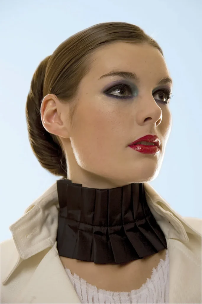 Een vrouw met achterovergekamd haar draagt opvallende make-up, met donkerblauwe oogschaduw en glanzende rode lippenstift. Ze heeft een geplooide zwarte choker en een wit kledingstuk met kraag. De achtergrond, geschikt voor bedrijfsfotografie, is een verloop van lichtblauw naar wit, waardoor een zachte, minimalistische setting ontstaat.