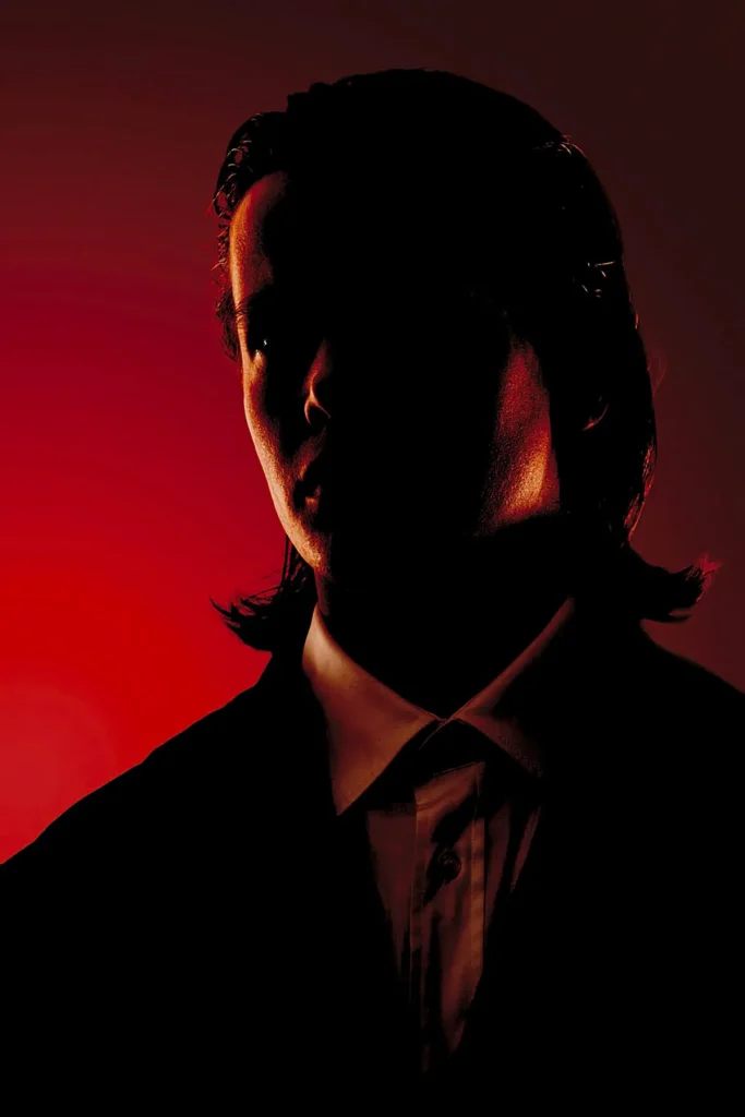 Een silhouetprofiel van een persoon met lang haar, gedeeltelijk verlicht, komt tegen een dramatische rode gradiëntachtergrond naar voren. Het individu draagt een jasje over een overhemd met kraag, wat een sfeer van mysterie en intriges uitstraalt die doet denken aan high-end bedrijfsfotografie, geaccentueerd door het samenspel van licht en schaduw.