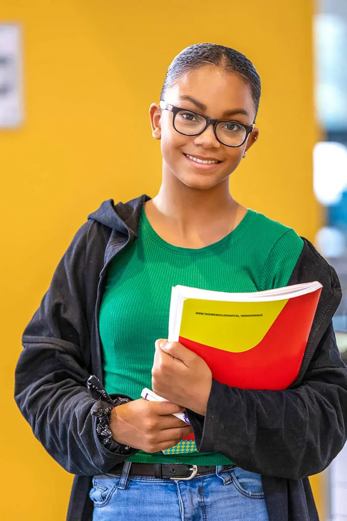 Een jongere met een bril en achterovergetrokken haar glimlacht terwijl hij een rood-gele map vasthoudt. Ze dragen een groen topje, een blauwe spijkerbroek en een zwart jasje tegen een felgele achtergrond. Dit beeld straalt vertrouwen en benaderbaarheid uit en geeft perfect de essentie van bedrijfsfotografie weer.