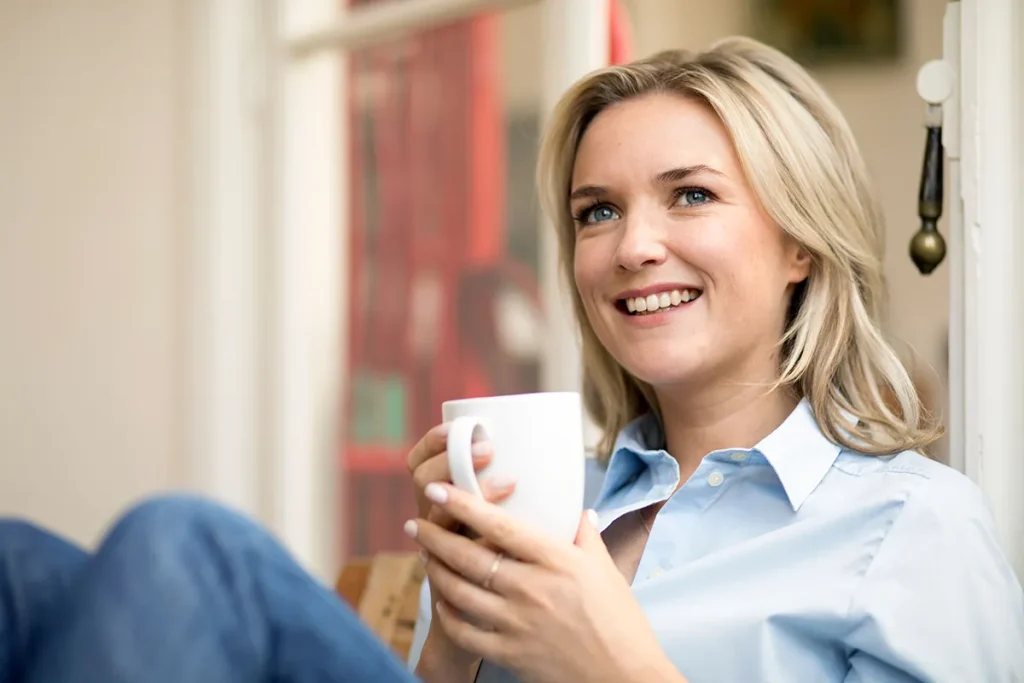 Een blonde vrouw in een lichtblauw shirt zit en glimlacht terwijl ze een witte mok met beide handen vasthoudt. Ze kijkt opzij en ziet er ontspannen en gelukkig uit. De setting, die doet denken aan professionele bedrijfsfotografie, bestaat uit een zacht vervaagde achtergrond met binnenelementen als een raam en een rood item.