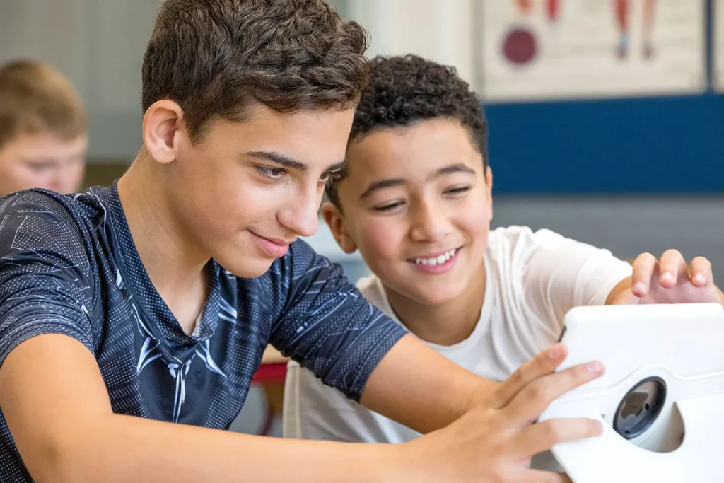 Twee jongens glimlachen en kijken naar een witte tablet die ze bij elkaar houden. De jongen links heeft kort, golvend, donker haar en draagt een overhemd met een donker patroon. De jongen rechts heeft kort krullend haar en draagt een wit overhemd. Ze zitten in een klaslokaal met een andere jongen wazig op de achtergrond, waardoor een perfect moment van bedrijfsfotografie wordt vastgelegd.