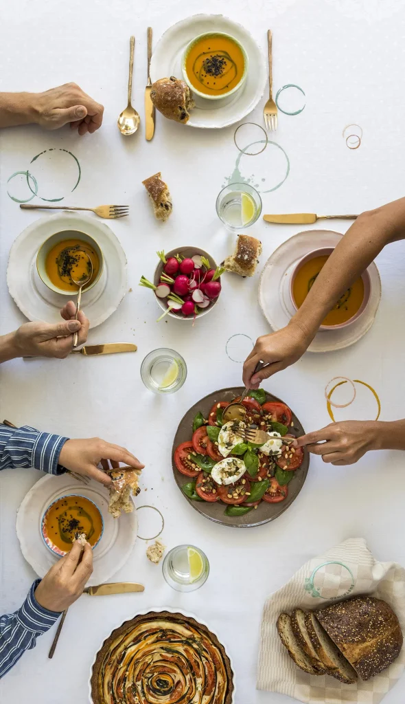Een bovenaanzicht van een tafel die gedekt is voor een maaltijd. Vier handen reiken naar verschillende gerechten, waaronder kommen soep, een bord radijsjes, een salade van tomaten en mozzarella en een groentetaart. Ook drankjes en sneetjes brood zijn aanwezig, samen met gouden bestek en witte borden op een wit tafelkleed: perfect voor bedrijfsfotografie.