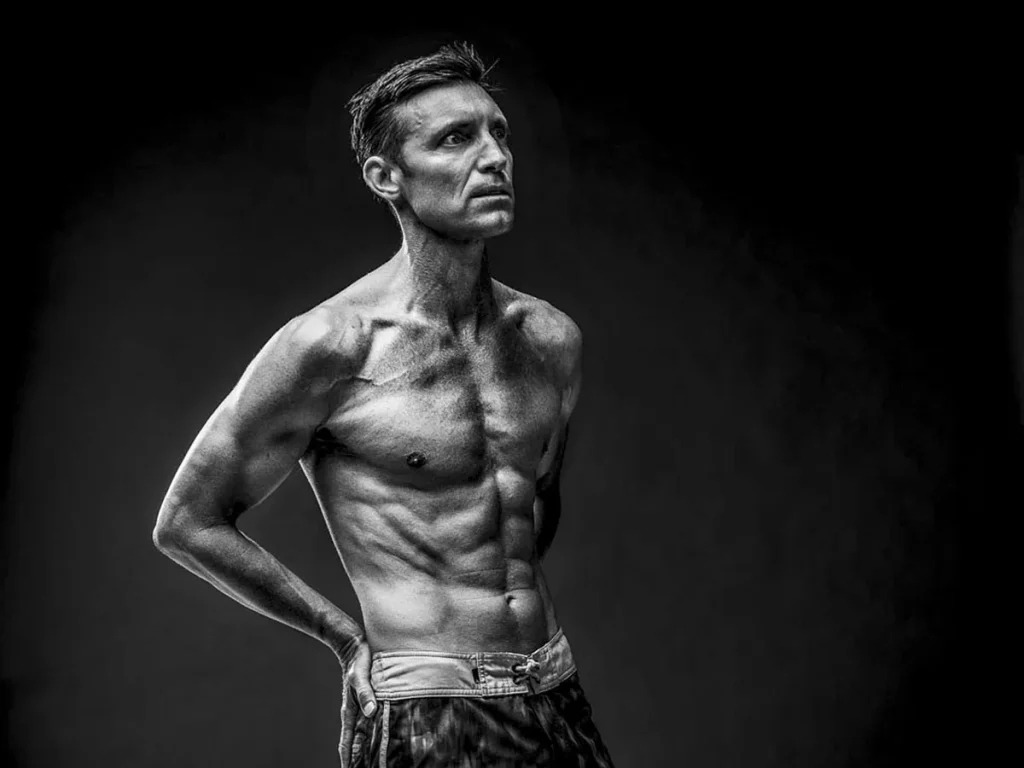 In een zwart-wit beeld poseert een shirtloze, gespierde man tegen een effen donkere achtergrond. Met kort haar en zichtbare buikspieren houdt hij zijn handen achter zijn rug en kijkt contemplatief naar boven en naar links. Zijn strakke lichaam, gedefinieerd en atletisch, straalt de precisie van bedrijfsfotografie uit.
