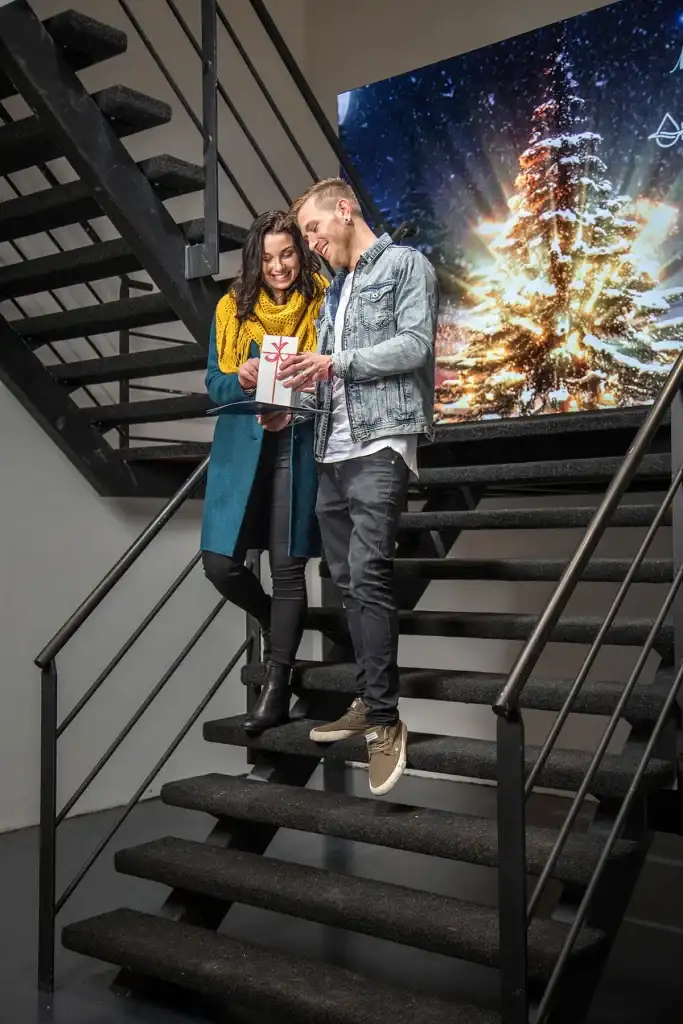 Een glimlachende vrouw en man staan op een trap, terwijl ze een ingepakt cadeau vasthouden en bekijken. De vrouw draagt een blauwgroen jas en een gele sjaal, terwijl de man een spijkerjasje draagt. Daarachter staat een groot scherm met een verlichte kerstboom in een besneeuwd nachttafereel, perfect voor bedrijfsfotografie.