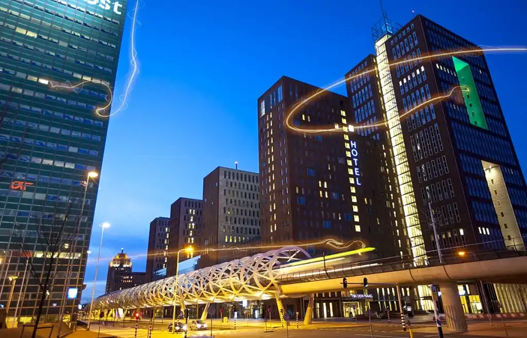 Een nachtelijk stadsbeeld met hoge moderne gebouwen, waarvan sommige het opschrift 'Hotel' hebben, verlicht tegen een helderblauwe lucht. Op de voorgrond steekt een helder verlichte, architectonisch ingewikkelde voetgangersbrug een weg over met lichte sporen van autoverkeer. Deze scène vat de essentie van stedelijke dynamiek en bedrijfsfotografie samen.