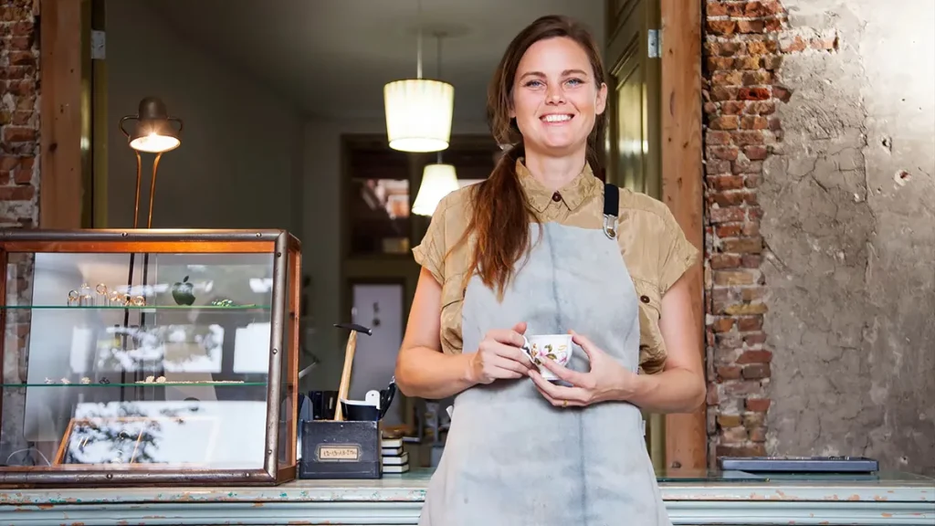 Een vrouw in een beige shirt en grijs schort glimlacht terwijl ze een kopje vasthoudt en achter een toonbank staat. De achtergrond bestaat uit bakstenen muren en hangende lampen, met een glazen vitrine waarin kleine voorwerpen worden tentoongesteld. Dit prachtig vastgelegde tafereel zou makkelijk te zien zijn in bedrijfsfotografie voor een ambachtswinkel of café.
