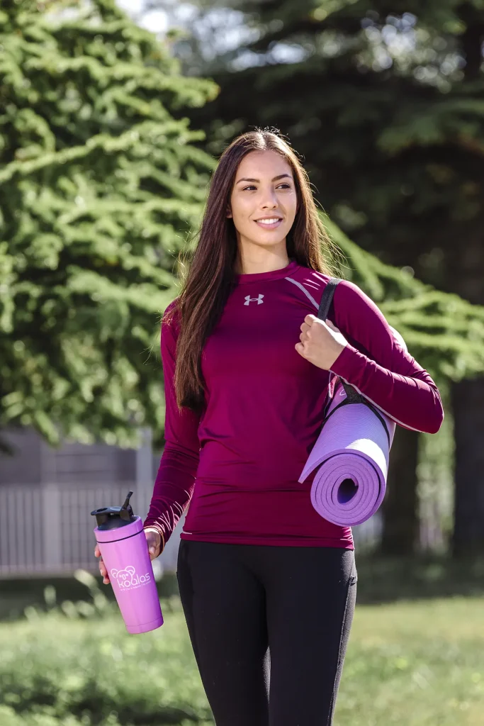 Een vrouw in sportkleding staat buiten met een paarse yogamat over haar schouder en een beker met het "ProBros"-logo in haar hand. Ze draagt een kastanjebruin topje met lange mouwen en een zwarte legging, met bomen en een gebouw op de achtergrond. Deze scène zou goed kunnen dienen voor bedrijfsfotografie, aangezien ze klaar lijkt voor een training.