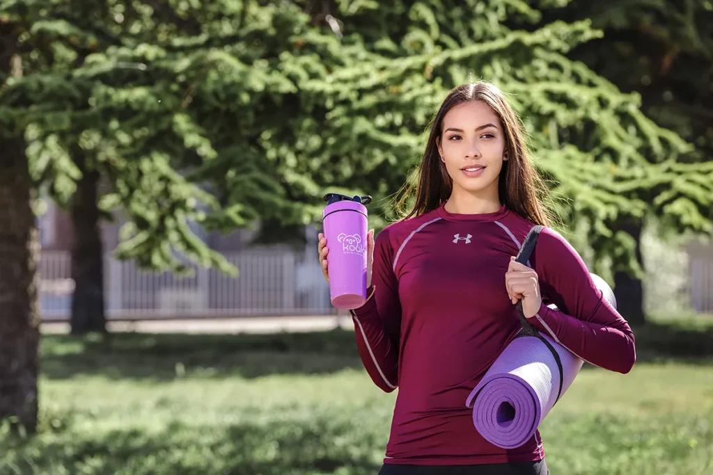 Een vrouw met lang donker haar, gekleed in een kastanjebruin sportshirt met lange mouwen, staat buiten met een paarse waterfles en een opgerolde paarse yogamat. Ze heeft de yogamat met een riem over haar schouder gehangen. Op de achtergrond zijn bomen en een hek zichtbaar, waardoor een ideaal bedrijfsfotografiemoment wordt vastgelegd.