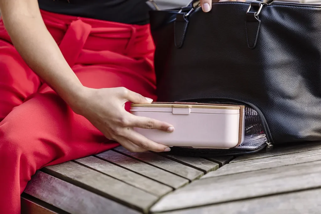 Een persoon die een rode broek en een zwart topje draagt, plaatst een lichtroze rechthoekige container in een zwartleren handtas. De persoon zit op een houten bank en de handtas is gedeeltelijk open, alsof hij bezig is met het ordenen van spullen: een ideaal tafereel voor bedrijfsfotografie.