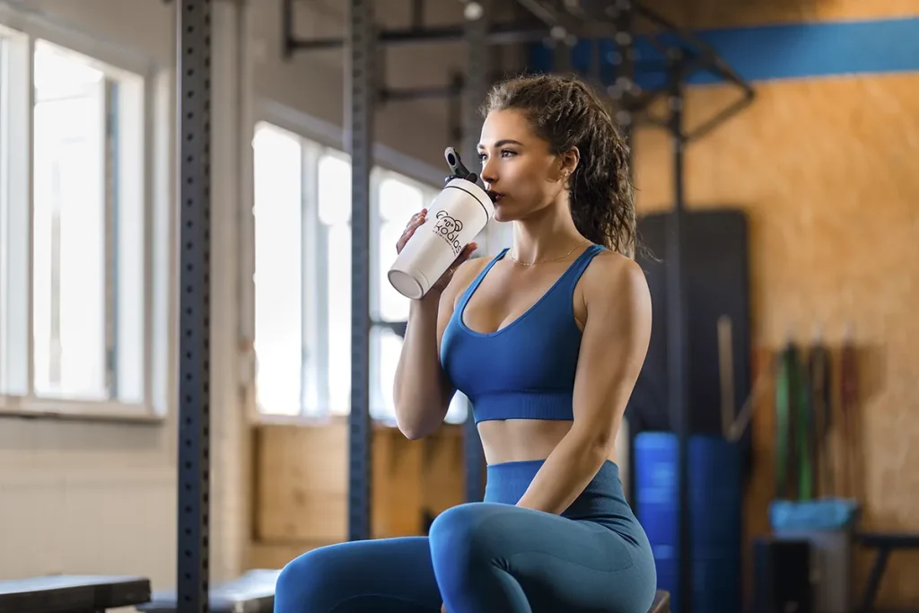 Een vrouw in blauwe sportkleding zit op een sportschoolbank en nipt van een eiwitshakerfles. Ze bevindt zich in een goed verlichte sportschool met grote ramen, zichtbare balken en diverse fitnessapparatuur op de achtergrond. De scène geeft de essentie van bedrijfsfotografie weer, terwijl het lijkt alsof ze een pauze neemt van haar trainingsroutine.