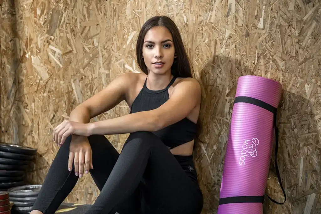 Een vrouw met lang bruin haar zit op de vloer tegen een gestructureerde houten muur. Ze draagt een zwart mouwloos topje en een zwarte legging. Naast haar ligt een opgerolde roze yogamat. Op de achtergrond zijn in de hoek halterschijven gestapeld, waardoor een element van bedrijfsfotografie wordt benadrukt. Ze kijkt rustig naar de camera.