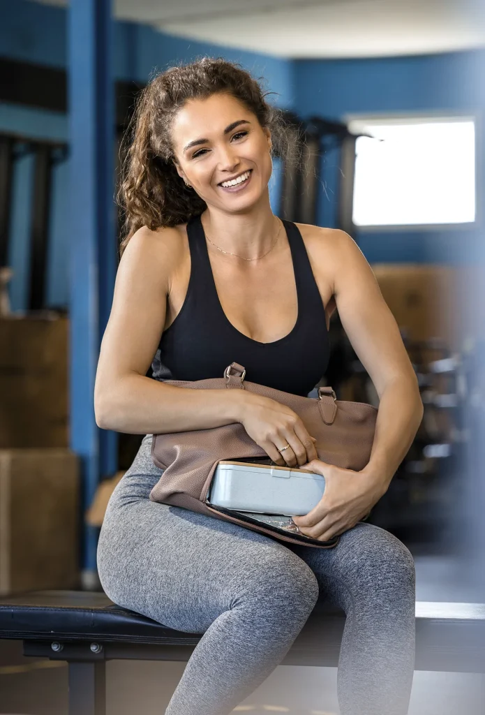 Een vrouw met bruin krullend haar in een paardenstaart zit op een bankje in een sportschool. Ze draagt een zwarte tanktop en een grijze legging en lacht vrolijk terwijl ze een bruine sporttas en een zilveren waterfles vasthoudt. De achtergrond van de sportschool, perfect voor bedrijfsfotografie, is voorzien van trainingsapparatuur en zacht, diffuus natuurlijk licht.