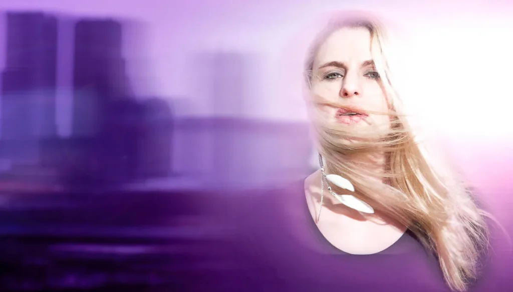 Een vrouw met lang blond haar staat voor een wazige stadsachtergrond, badend in paars licht. Haar haar wappert over haar gezicht en ze draagt een zwart topje en een grote, bungelende oorbel. Het totaalbeeld heeft een dromerige, etherische kwaliteit die doet denken aan high-end bedrijfsfotografie.
