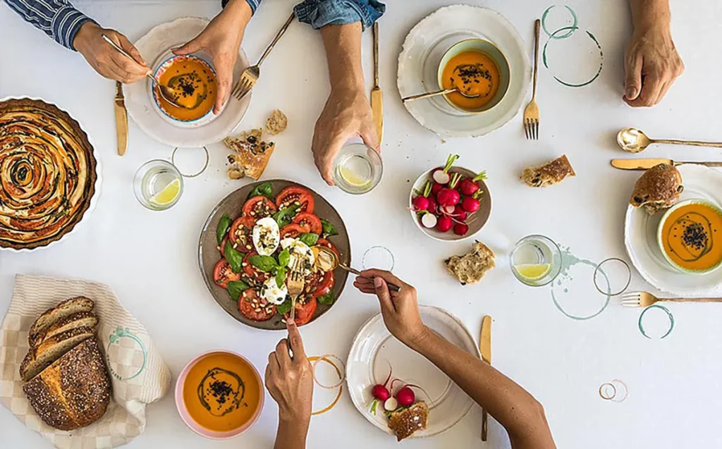 Een bovenaanzicht van een maaltijd waarbij de handen van drie mensen naar het eten reiken, is een perfect moment voor bedrijfsfotografie. Op de tafel staan kommen sinaasappelsoep, gesneden brood, een kleurrijke tomatensalade met mozzarella en basilicum, een taart en radijsjes. Drankjes in heldere glazen liggen verspreid over het witte tafelkleed.
