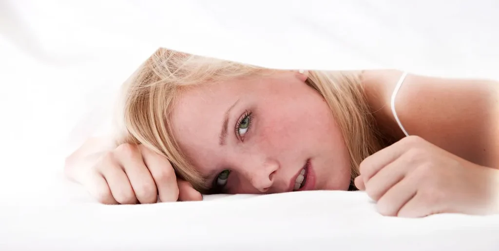 Een persoon met blond haar gluurt onder witte lakens vandaan, zijn hoofd rust op een wit oppervlak en kijkt met een kalme uitdrukking rechtstreeks naar de camera. Beide handen zijn zichtbaar en houden de lakens boven hun hoofd. De overwegend witte achtergrond zorgt voor een zachte, serene sfeer die doet denken aan bedrijfsfotografie.