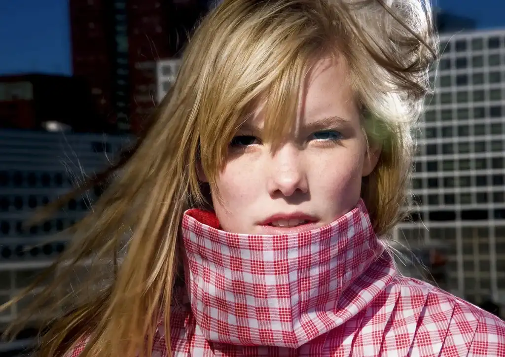 Een vrouw staart recht in de camera, haar blonde haar wappert in de wind. Ze draagt een roze en wit geruit jasje met hoge kraag dat haar mond gedeeltelijk bedekt, wat een professionele look belichaamt die perfect is voor bedrijfsfotografie. Onduidelijke hoge gebouwen doemen op de achtergrond op onder een helderblauwe hemel.
