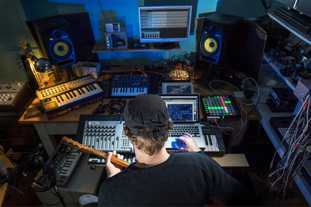 Een persoon met een zwarte pet op zit in een rommelige muziekproductieopstelling met meerdere monitoren, toetsenborden, een drumpad en diverse audioapparatuur. Ze houden een gitaar vast en gebruiken een computermuis, met schermen waarop muzieksoftware wordt weergegeven: de perfecte scène voor bedrijfsfotografie.