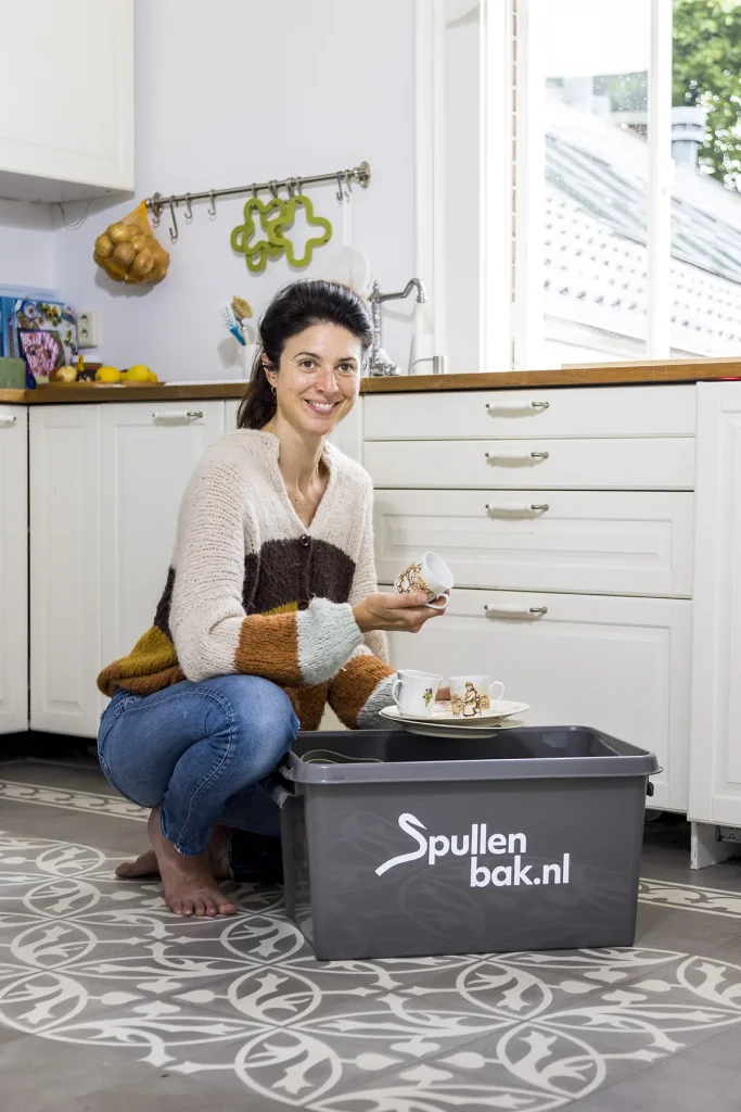 Een vrouw hurkt in een keuken, met een theekop en schotel in haar hand. Ze heeft donker haar dat naar achteren is gebonden en draagt een gezellige trui met kleurblokken en een blauwe spijkerbroek. Voor haar staat een grijze plastic bak met de tekst 'Spullenbak.nl', waarmee een alledaags moment wordt vastgelegd, perfect voor bedrijfsfotografie. De keuken heeft witte kasten en tegelvloeren met patronen.