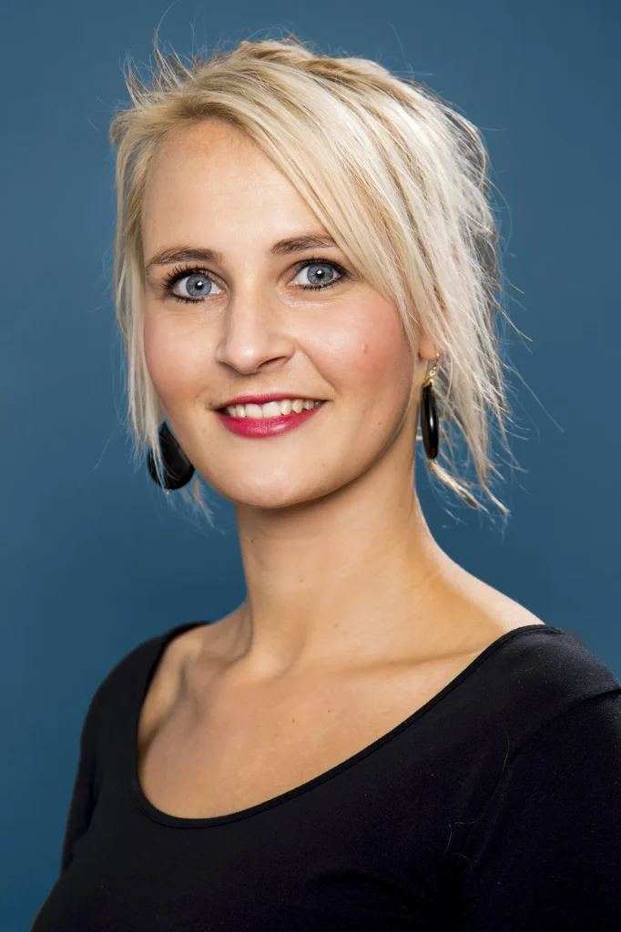 Een vrouw met blond haar, losjes opgestoken, lacht naar de camera. Ze heeft felblauwe ogen, draagt rode lippenstift en heeft minimale make-up. Gekleed in een zwart topje en versierd met zwarte ronde oorbellen, staat ze tegen een effen donkerblauwe achtergrond: een perfect voorbeeld van bedrijfsfotografie.