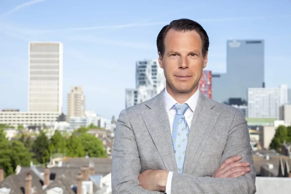 Een man in een lichtgrijs pak met een lichtblauw overhemd en een stropdas met een groenblauw patroon staat met de armen over elkaar geslagen. Hij is buiten met op de achtergrond een stadsgezicht met diverse moderne gebouwen en groen. De lucht is helder en blauw, waardoor een ideaal tafereel ontstaat voor bedrijfsfotografie.