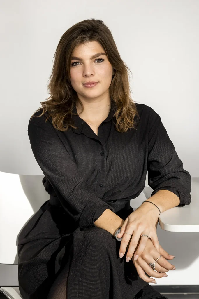 Een persoon met schouderlang, golvend bruin haar zit in een moderne, minimalistische stoel. Ze dragen een zwarte jurk met lange mouwen en knopen en hebben hun handen op hun schoot, waar verschillende ringen zichtbaar zijn. De achtergrond is eenvoudig en licht, waardoor de focus op het individu wordt versterkt: perfect voor bedrijfsfotografie.