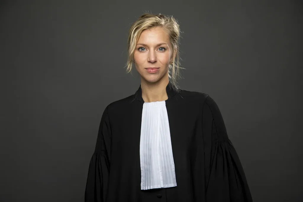 Tegen een donkergrijze achtergrond staat een vrouw, gekleed in een traditioneel advocatengewaad. Het gewaad is zwart met wijde mouwen en ze heeft een witte nekband, typisch voor rechterlijke kledij. Met blond haar losjes gestyled en met een neutrale uitdrukking rechtstreeks in de camera kijkend, belichaamt dit beeld professionele bedrijfsfotografie.