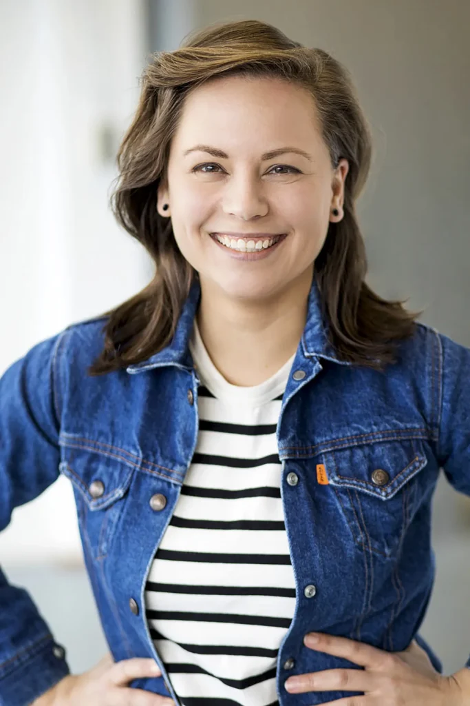 Een glimlachende persoon met bruin haar op schouderlengte staat met de handen op de heupen. Ze dragen een blauw spijkerjack over een zwart-wit gestreept overhemd. De achtergrond, zacht verlicht en onscherp, creëert een neutrale binnenomgeving die professionaliteit uitstraalt, perfect voor bedrijfsfotografie.