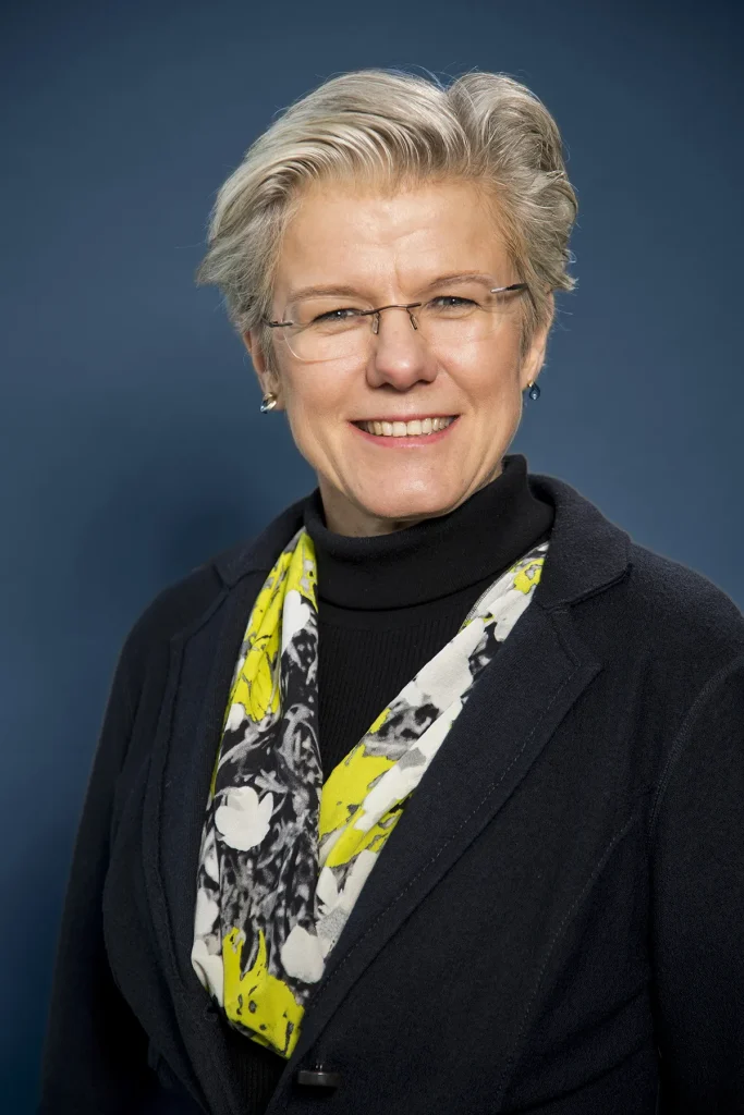 Een persoon met kort grijs haar en een bril glimlacht, gekleed in een donkere blazer over een zwarte coltrui. Ze dragen een witte sjaal met gele en zwarte patronen. De achtergrond, ideaal voor bedrijfsfotografie, is effen donkerblauw.