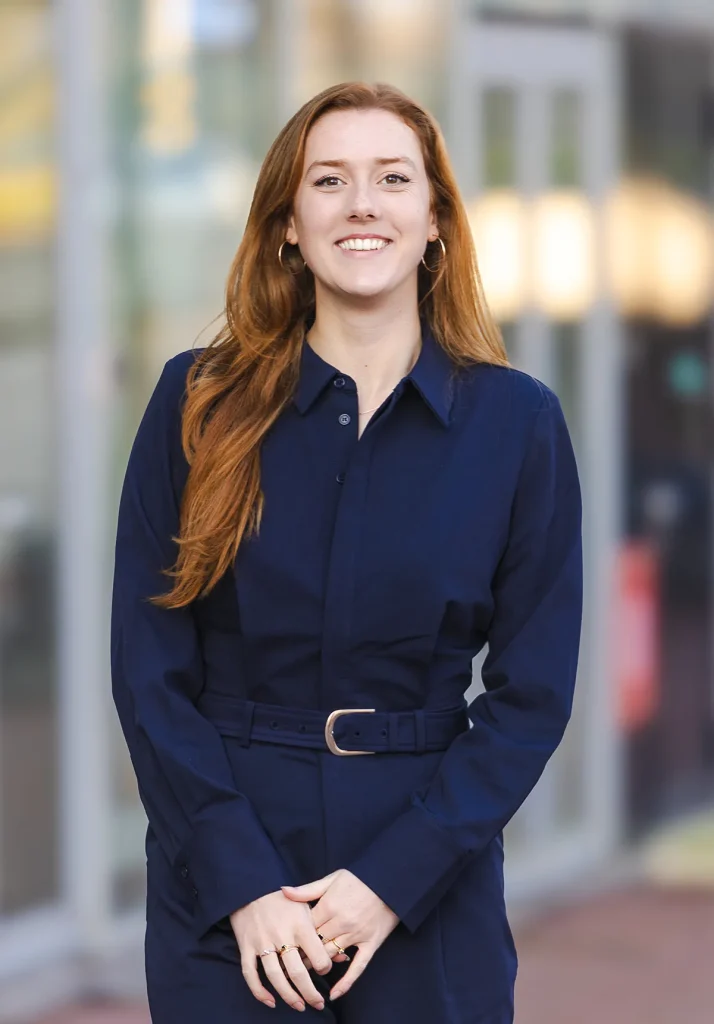 Een vrouw met lang rood haar staat glimlachend buiten, wat een perfect moment van bedrijfsfotografie belichaamt. Ze draagt een donkerblauwe jurk met lange mouwen en een riem. De achtergrond is wazig en toont een gebouw met grote ramen en diffuus licht, wat duidt op een stedelijke omgeving.
