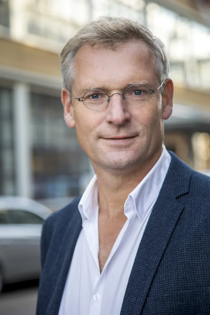 Een man met kort, licht haar en een bril staat buiten. Hij draagt een wit overhemd, met een iets losgeknoopte kraag, onder een donkerblauwe blazer. De achtergrond, vervaagd in professionele bedrijfsfotografiestijl, toont een gebouw met grote ramen en een geparkeerde auto. Hij heeft een serieuze uitdrukking en kijkt rechtstreeks in de camera.