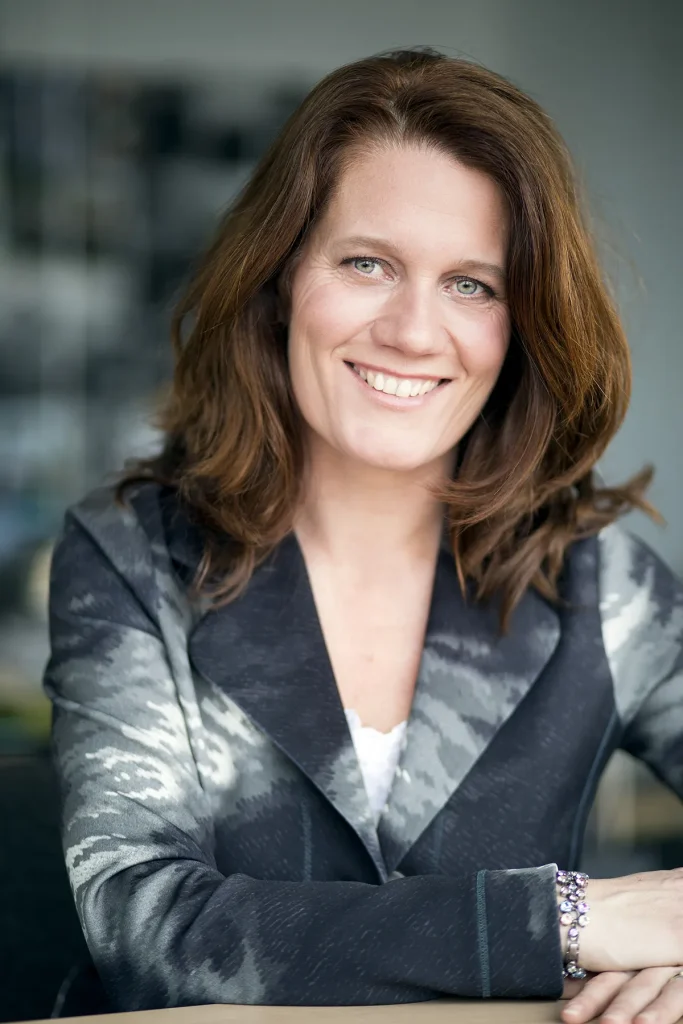 Een vrouw met schouderlang bruin haar lacht hartelijk naar de camera. Ze draagt een blazer met een patroon met grijstinten en een witte top eronder. Ze heeft lichtgekleurde ogen en zit tegen een onscherpe, niet te onderscheiden achtergrond, perfect voor bedrijfsfotografie. Haar handen rusten op een oppervlak voor haar.