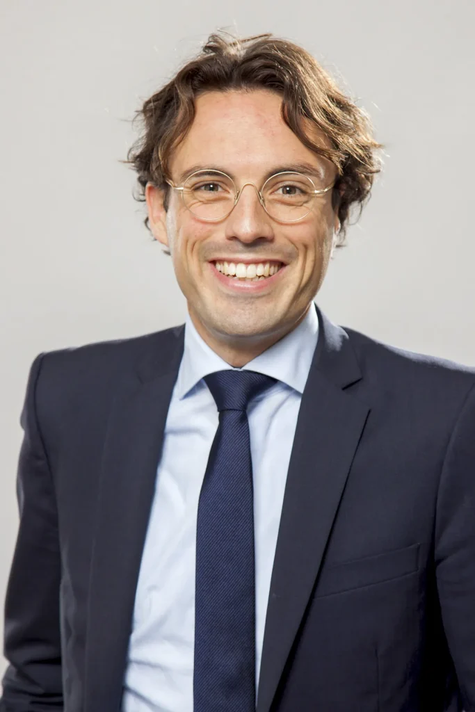 Een persoon met golvend bruin haar en een bril glimlacht. Ze dragen een donkerblauw colbert, een lichtblauw overhemd en een donkerblauwe stropdas. De achtergrond is effen en lichtgekleurd en geeft met zijn professionele en toch toegankelijke uitstraling de essentie van bedrijfsfotografie weer.