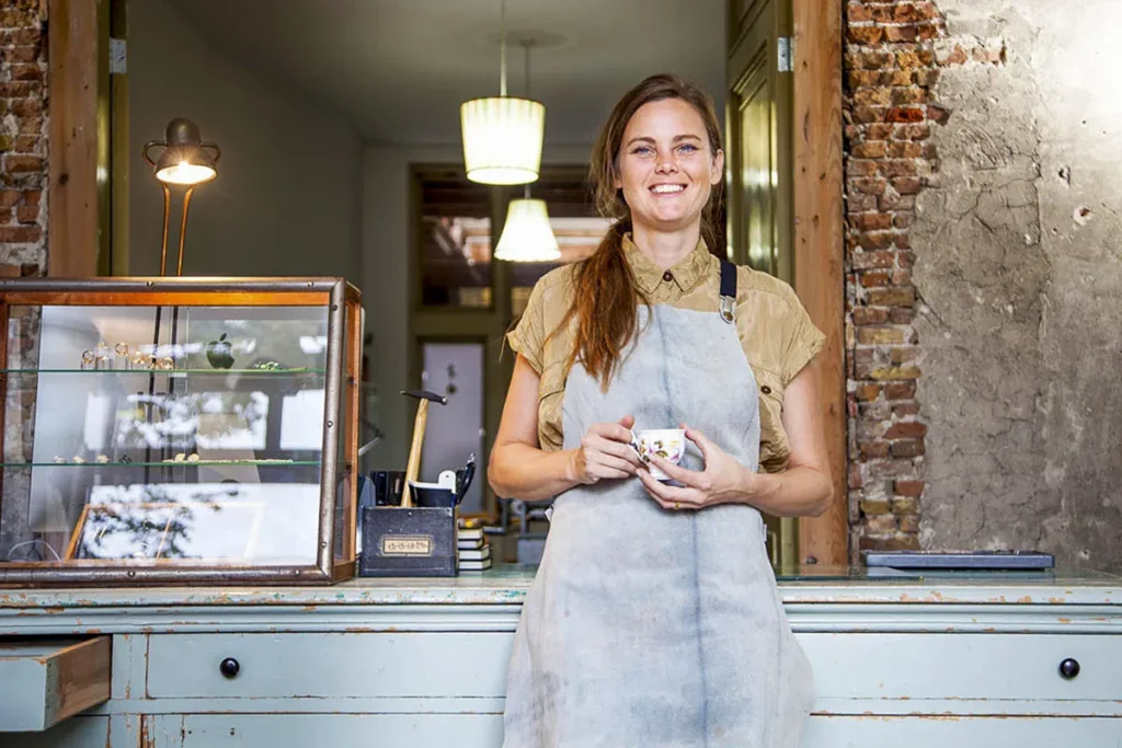 Een glimlachende vrouw in een grijs schort staat achter een lichtblauwe toonbank in een rustieke winkel en belichaamt de essentie van bedrijfsfotografie. Ze houdt een klein, kleurrijk kopje vast. Achter haar staat een vitrine met diverse spullen en twee hanglampen. De winkel heeft bakstenen muren en een uitnodigende sfeer.