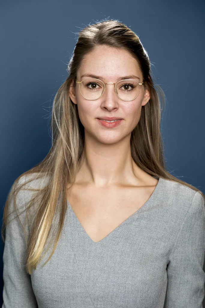 Een vrouw met lang blond haar en een bril glimlacht lichtjes terwijl ze een lichtgrijs topje met V-hals draagt. Ze staat tegen een gladde donkerblauwe achtergrond en kijkt recht naar de camera. Haar haar is netjes gescheiden en achter de oren gestopt, wat de professionele uitstraling belichaamt die ideaal is voor bedrijfsfotografie.