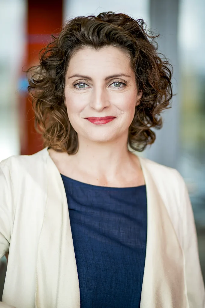 Een vrouw met schouderlang, bruin krullend haar lacht vriendelijk naar de camera en belichaamt de professionele elegantie die essentieel is voor bedrijfsfotografie. Ze draagt een marineblauw topje onder een lichtbeige blazer, tegen een zacht onscherpe achtergrond met hints van rode en blauwe tinten.