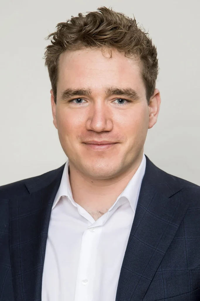 Een persoon met kort, golvend, lichtbruin haar en blauwe ogen wordt getoond in een hoofd-en-schouders bedrijfsfotografieportret. Ze dragen een donkerblauw pak en een wit overhemd, tegen een effen lichtgrijze achtergrond. Ze glimlachen lichtjes.