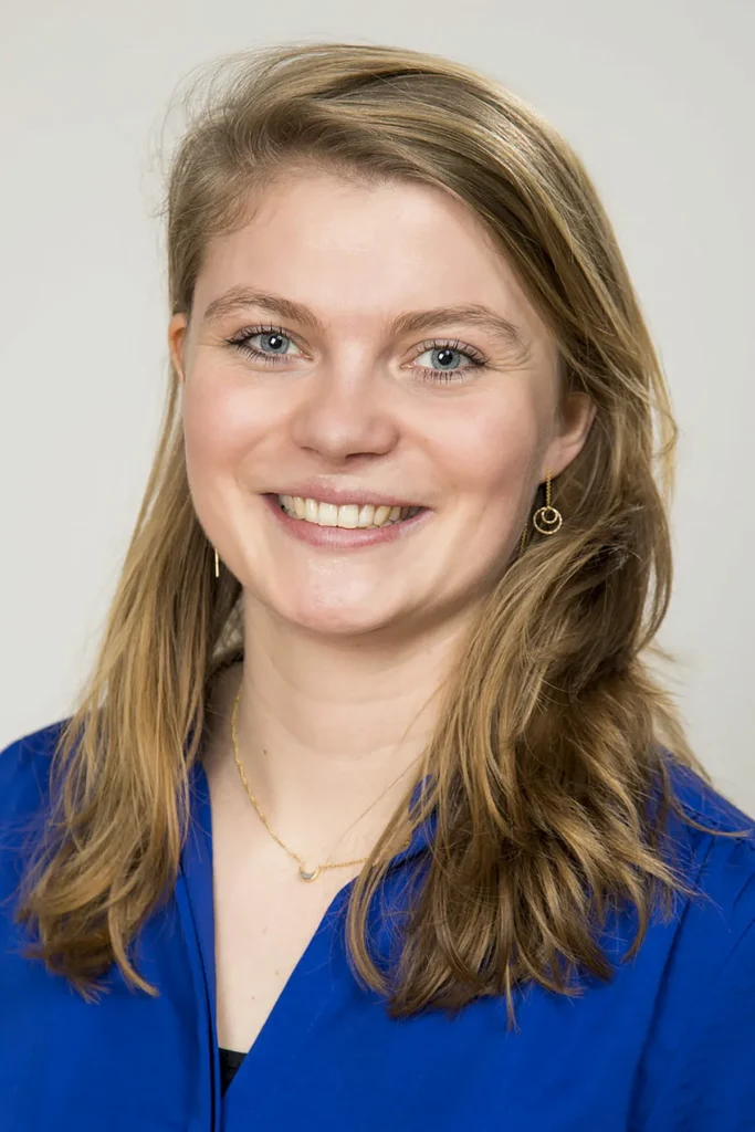 Een vrouw met lang, lichtbruin haar lacht naar de camera. Ze draagt een blauw topje en kleine gouden oorringen. Haar haar heeft een zijscheiding en valt over haar schouders. De achtergrond is effen en licht gekleurd, typisch voor bedrijfsfotografie, waardoor de focus op haar gezicht ligt.