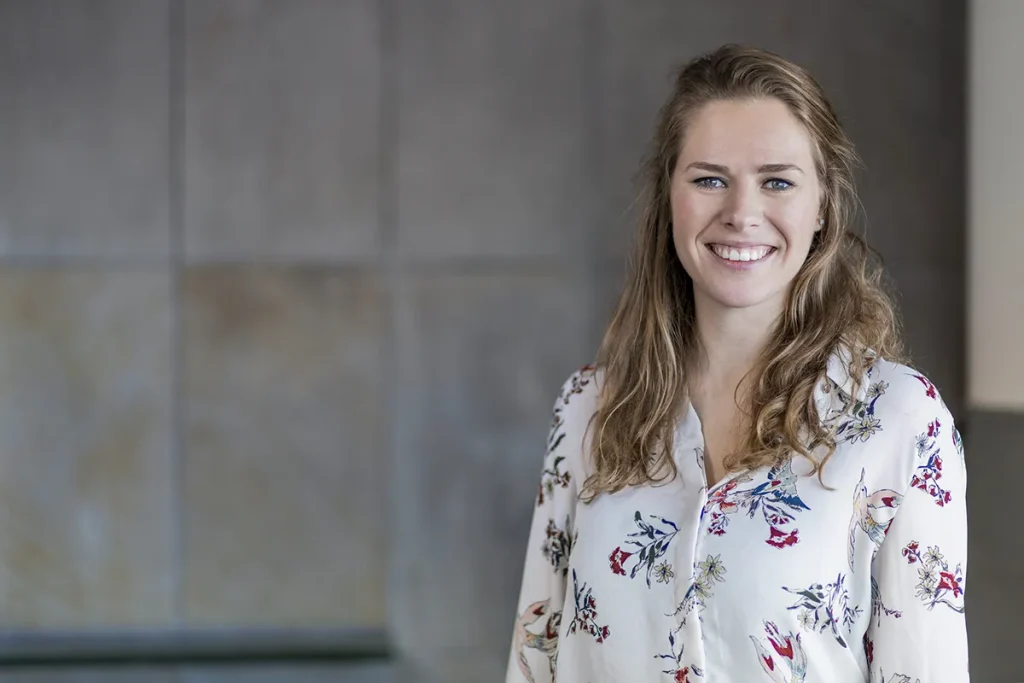 Een vrouw met lang golvend bruin haar glimlacht naar de camera. Ze draagt een witte blouse met een kleurrijk bloemenpatroon. De achtergrond is een wazige, neutraal getinte muur, waardoor het beeld een professionele en toch ontspannen sfeer krijgt, ideaal voor bedrijfsfotografie.
