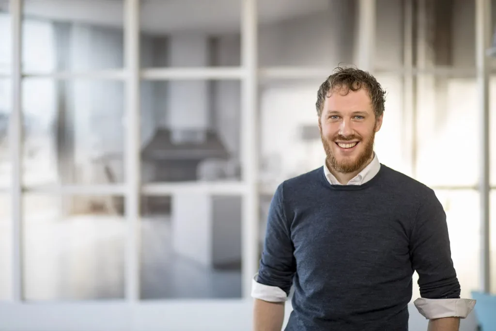 Een man met krullend haar en een baard, gekleed in een grijze trui over een overhemd met witte kraag, staat glimlachend in een kantooromgeving. De achtergrond bestaat uit grote glazen ramen en wazig kantoormeubilair, waardoor de essentie van moderne, professionele bedrijfsfotografie wordt vastgelegd.