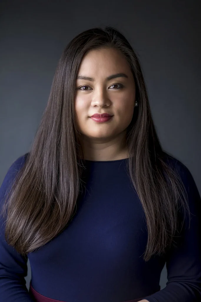 Een vrouw met lang, steil donker haar poseert tegen een donkergrijze achtergrond. Ze draagt een donkerblauw shirt en heeft een lichte, zelfverzekerde glimlach op haar gezicht. Haar lippen zijn dieprood geverfd en haar make-up is subtiel, met de nadruk op haar ogen. De zachte verlichting benadrukt haar gelaatstrekken, perfect voor bedrijfsfotografie.