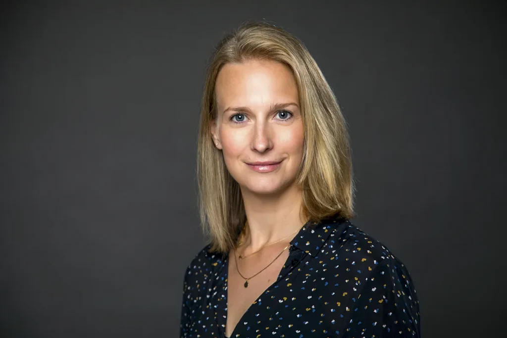 Een vrouw met blond haar en een blanke huid staat tegen een donkergrijze achtergrond. Ze draagt een donkere blouse met een subtiel patroon en een fijne ketting. Met een lichte glimlach en rustige uitdrukking belichaamt de vrouw professionaliteit, waardoor dit een ideaal voorbeeld van bedrijfsfotografie is.