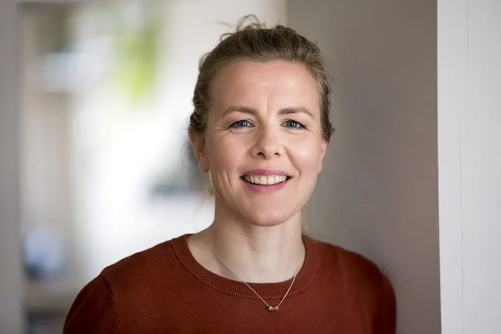 Een vrouw met een lichte huid en blond haar lacht hartelijk naar de camera. Ze draagt een roestkleurige trui en een klein, fijn halssnoer met hangertje. De achtergrond, typisch voor bedrijfsfotografie, is zacht vervaagd, wat een binnenomgeving met natuurlijk licht suggereert.