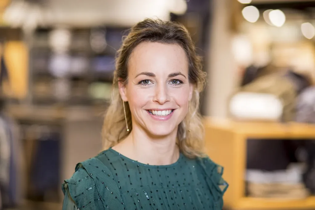 Een lachende vrouw met blond haar staat binnen. Ze draagt een groene blouse met ruches aan de mouwen en gouden oorbellen. De achtergrond is wazig, met planken en diverse spullen, wat een winkel- of bedrijfsfotografieomgeving suggereert. De verlichting is warm en uitnodigend.
