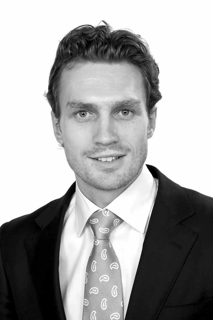 Een zwart-wit bedrijfsfotografieportret van een persoon met kort, golvend haar, gekleed in een donker pak, een wit overhemd en een paisley-das met patroon. De persoon lacht lichtjes en kijkt rechtstreeks in de camera tegen een effen witte achtergrond.